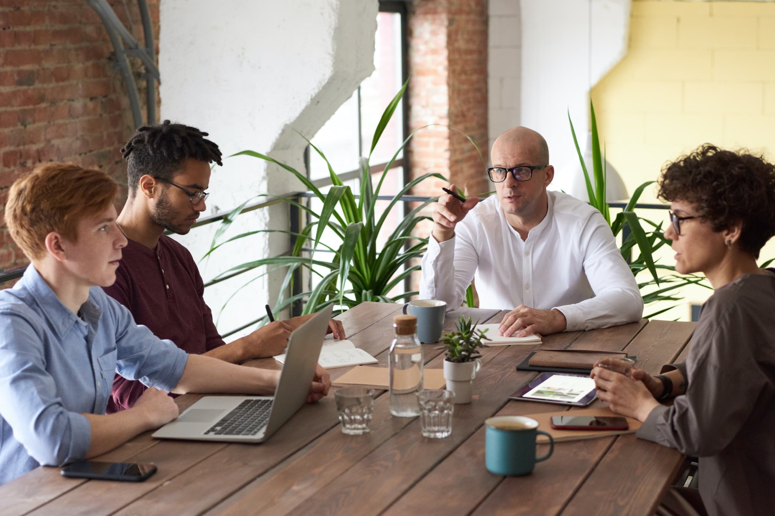 Team sitting around table, man sitting in the middle pointing to the group with his pen